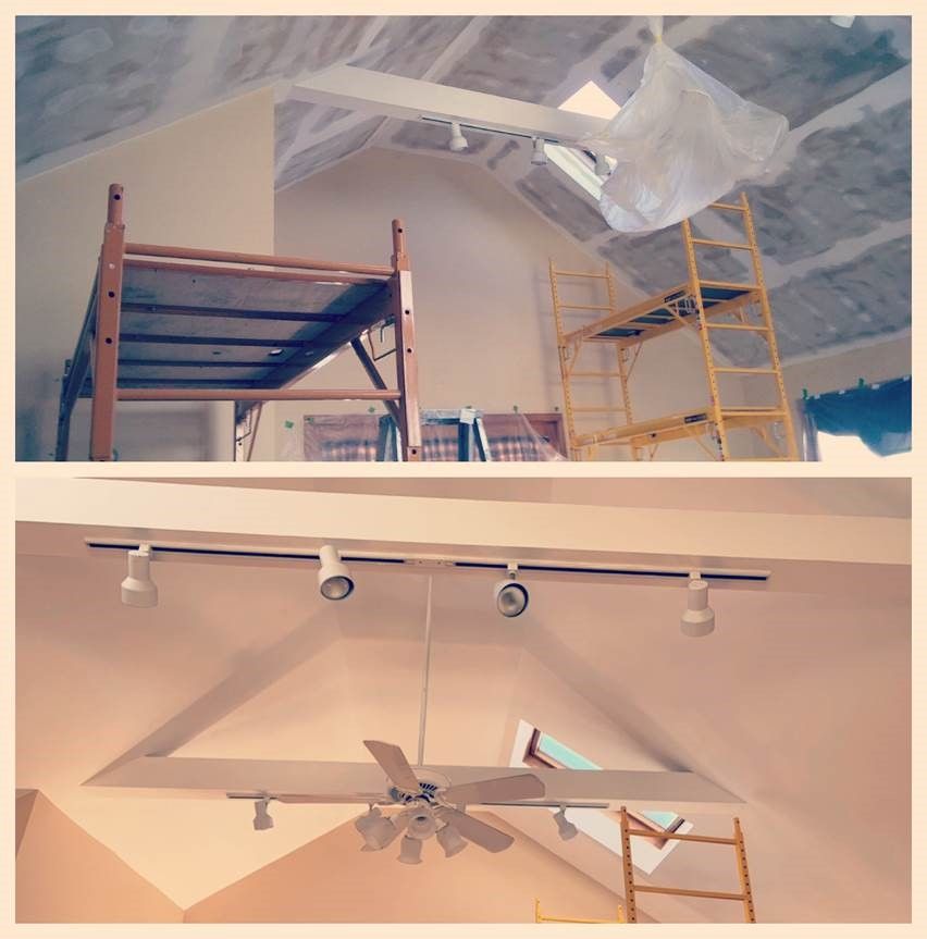 Scaffolding and a ladder in a room with vaulted ceiling being painted white; track lights and a ceiling fan below.