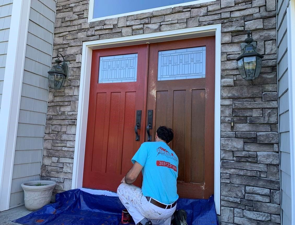 Person kneeling at a red double front door, painting the right door brown beside stone siding.