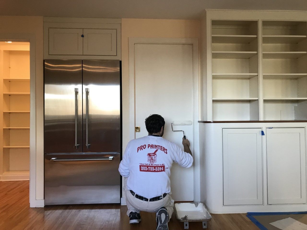Person painting a wall beside a stainless steel refrigerator in a bright kitchen