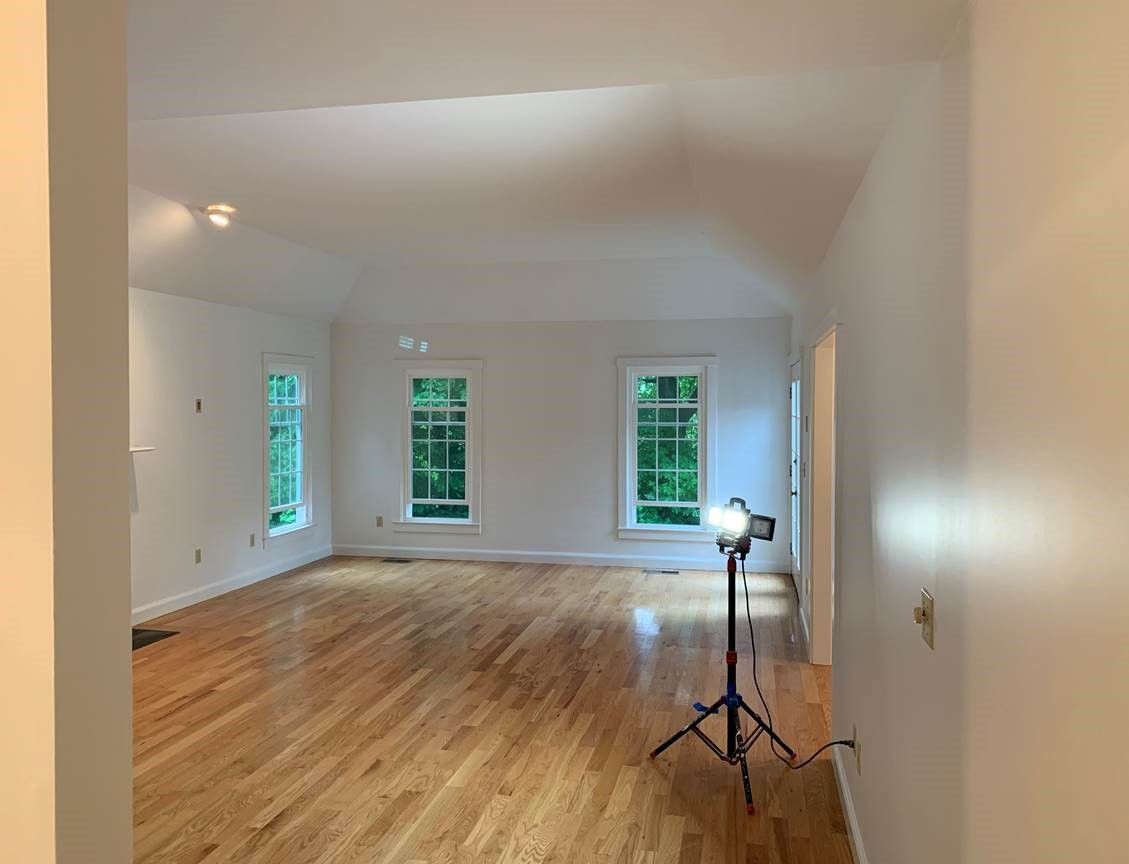 Empty white living room with hardwood floors and three windows, lit by a tripod work light