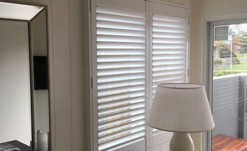 A Bedroom with White Shutters, a Lamp, a Mirror and A Sliding Glass Door — Phoenix Blinds In Albion Park Rail, NSW
