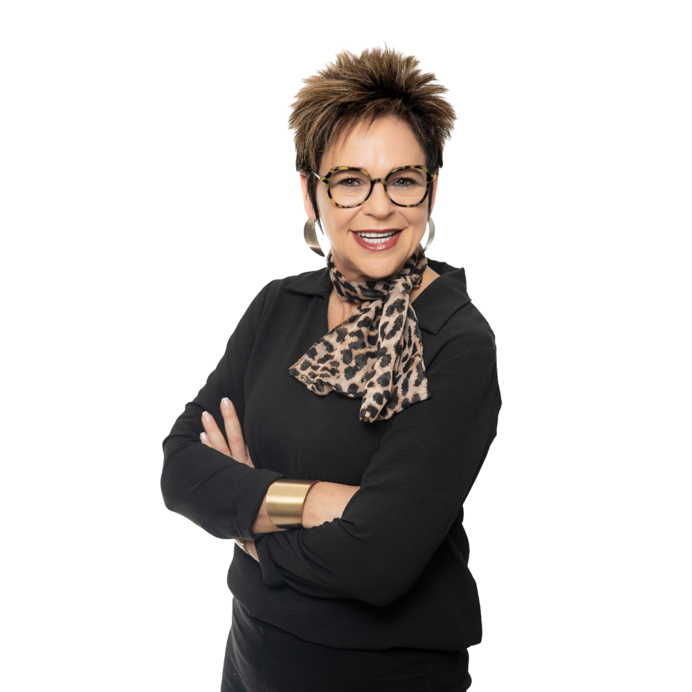 A person with short, dark, textured hair wears glasses, a leopard-print scarf, and a black top, smiling with arms crossed.