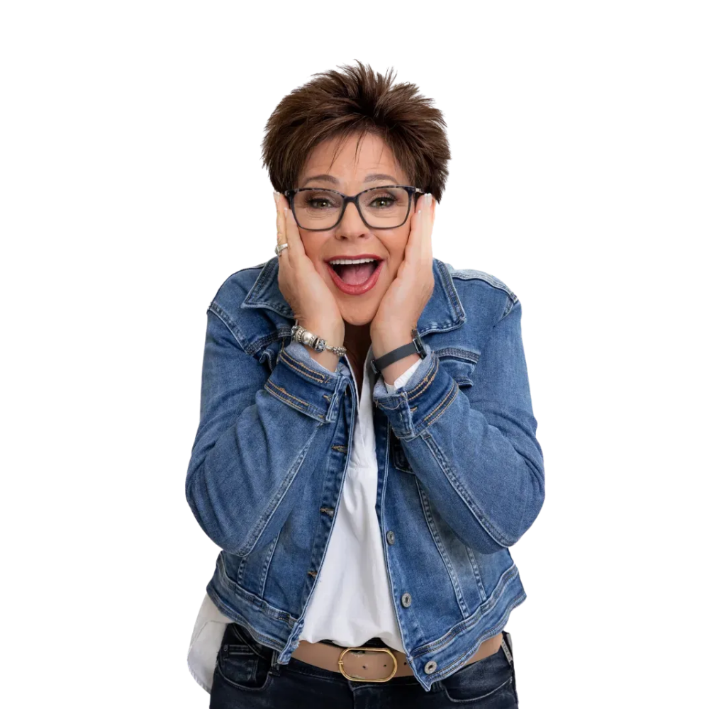 A person with short brown hair wearing glasses and a denim jacket, hands to their face, mouth open in an excited expression.