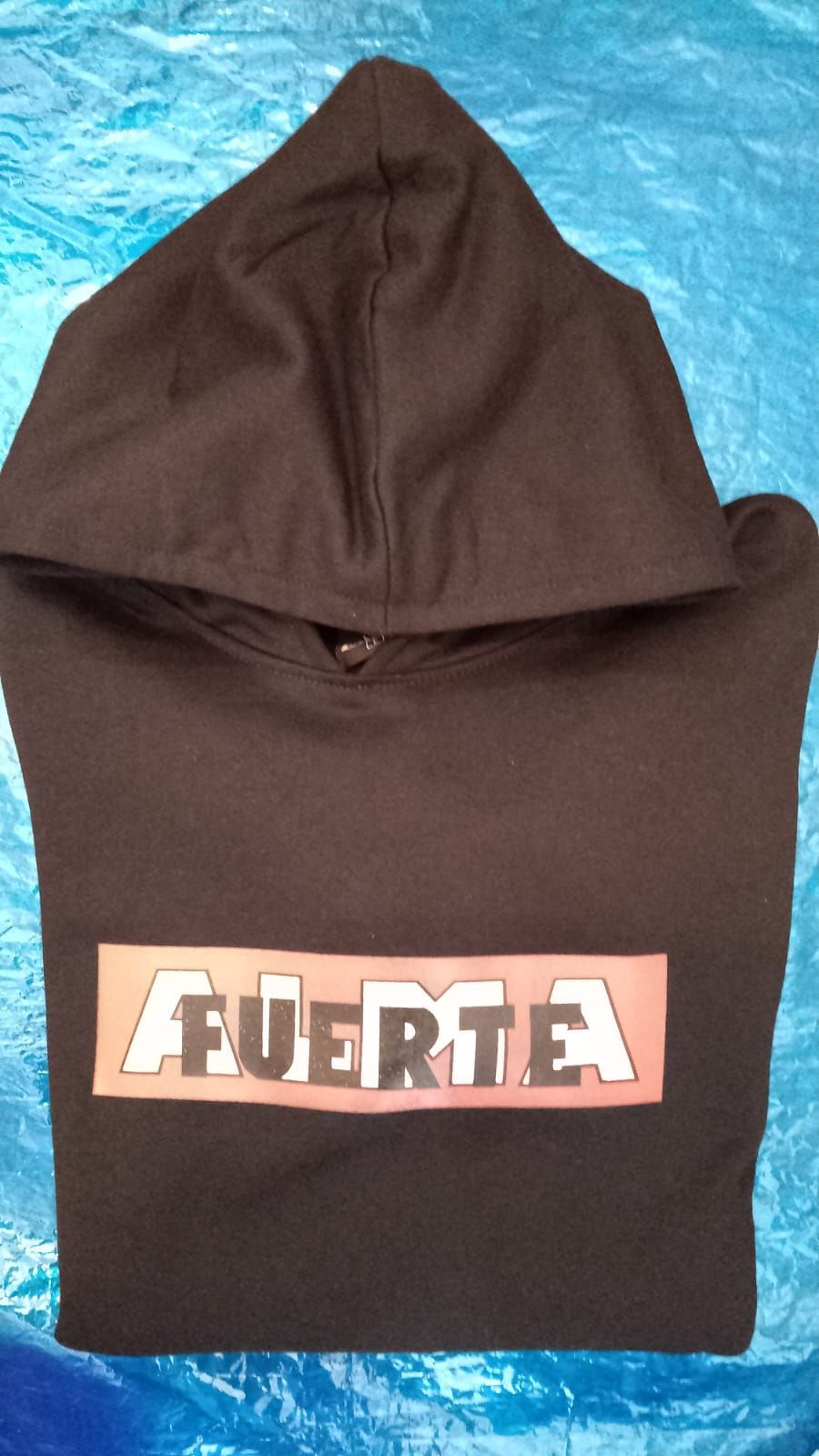 Sudadera marrón con capucha grande y el logotipo “FUERZA” en el pecho, sobre una superficie azul.
