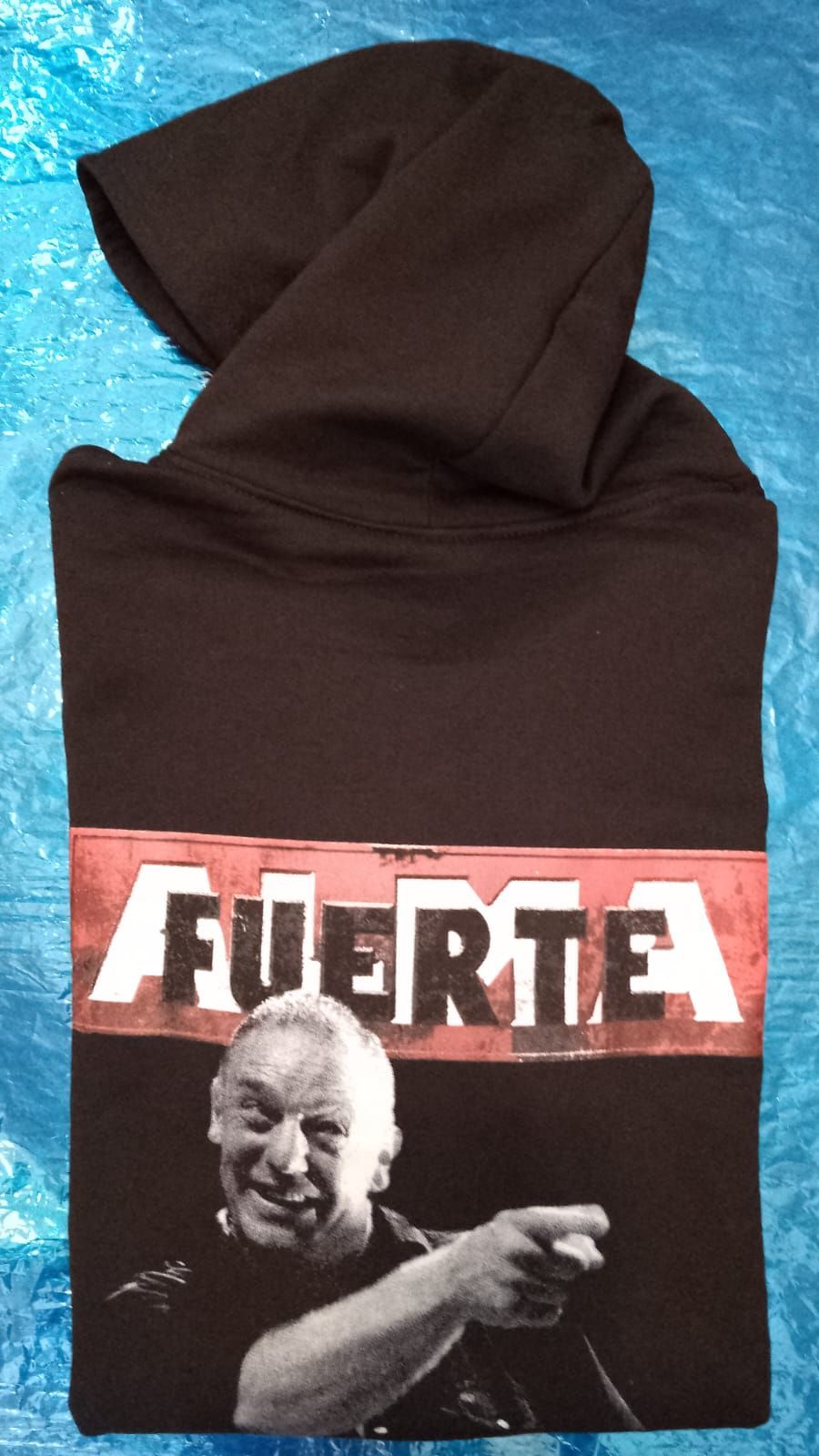 Sudadera marrón con capucha grande y el logotipo “FUERZA” en el pecho, sobre una superficie azul.