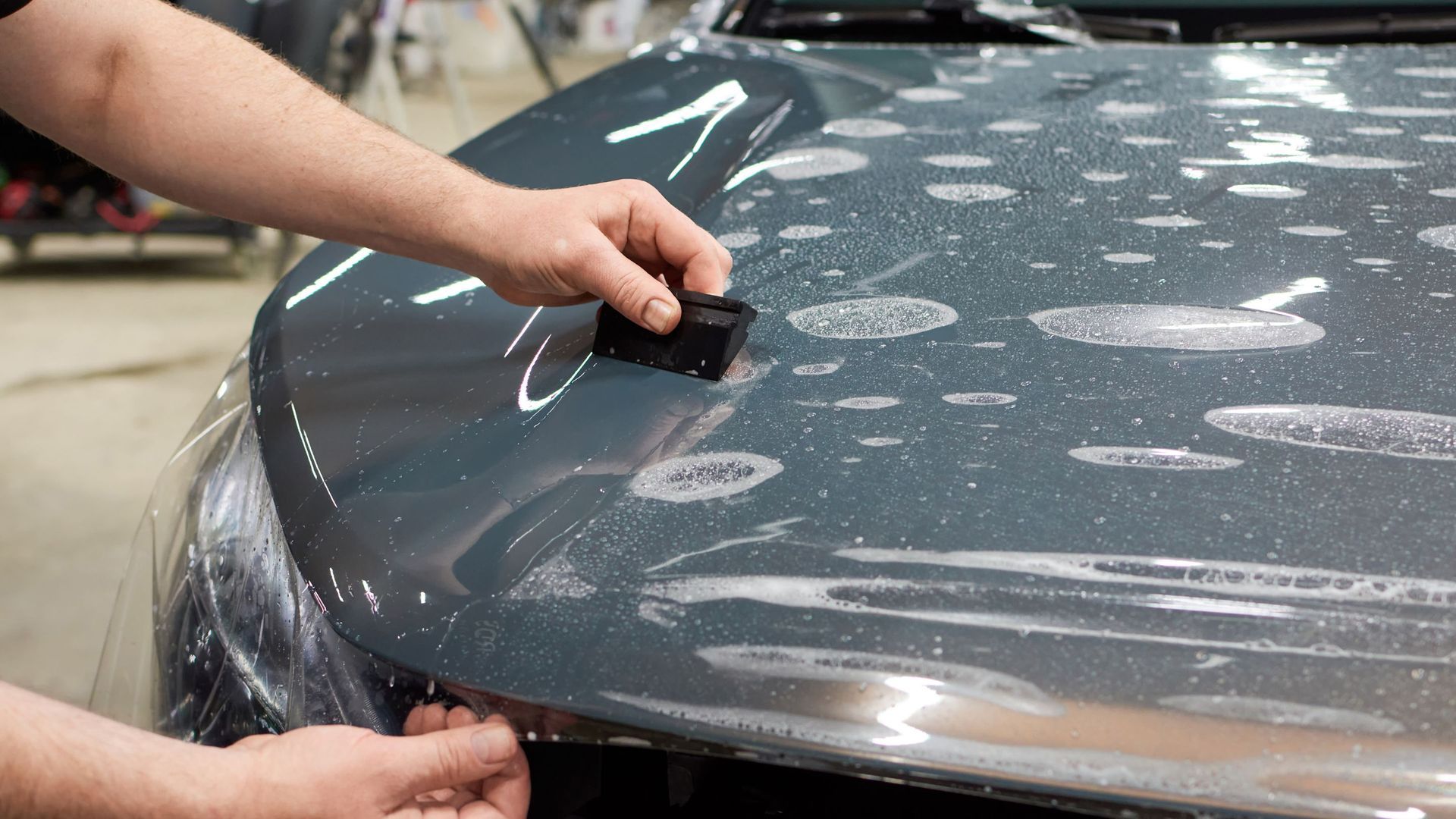 Person using a squeegee to apply a clear protective film to the hood of a car.