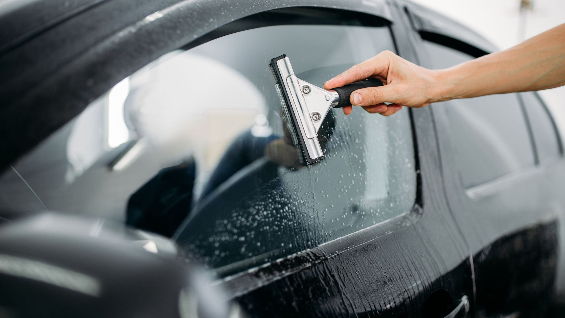 Person squeegeeing a car window, removing soapy water. Black car, bright interior.