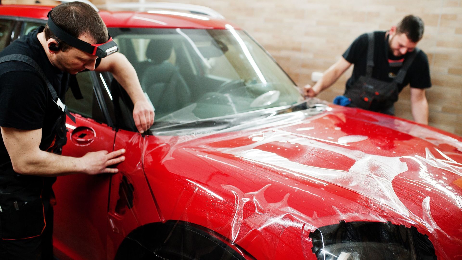 Two men installing clear film on a red car's front, in a garage.