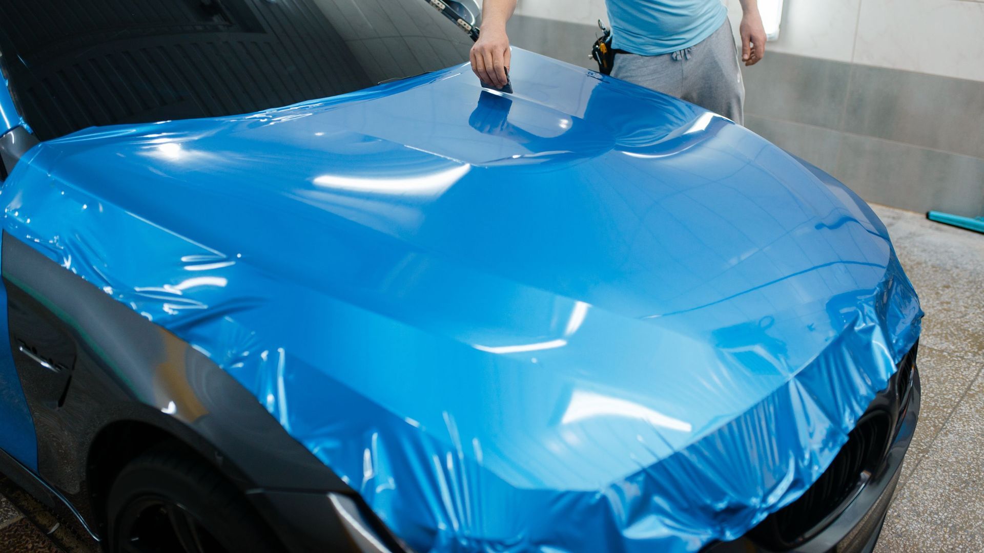 A person applying blue protective film to the hood of a car in a garage.