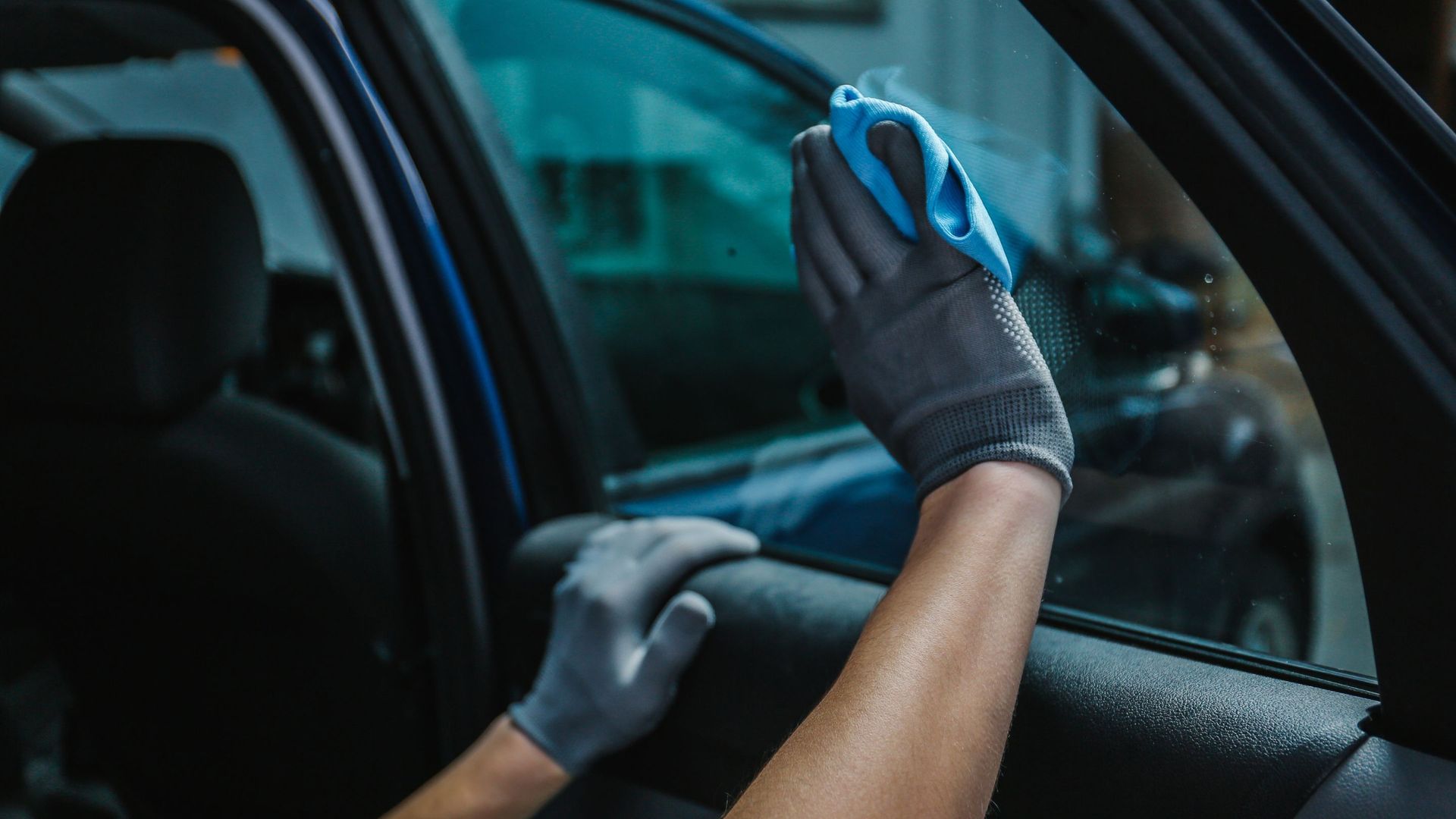 Person cleaning car window with blue cloth, wearing grey gloves.