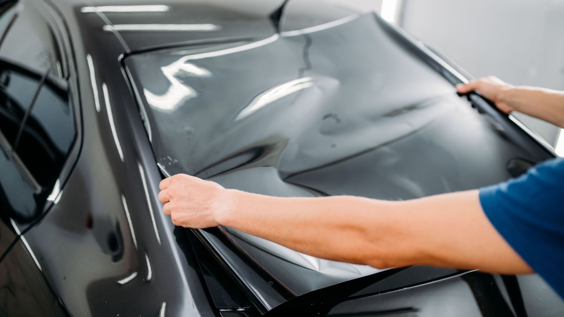 A person applying tinted film to a car's rear window with a squeegee in a garage.