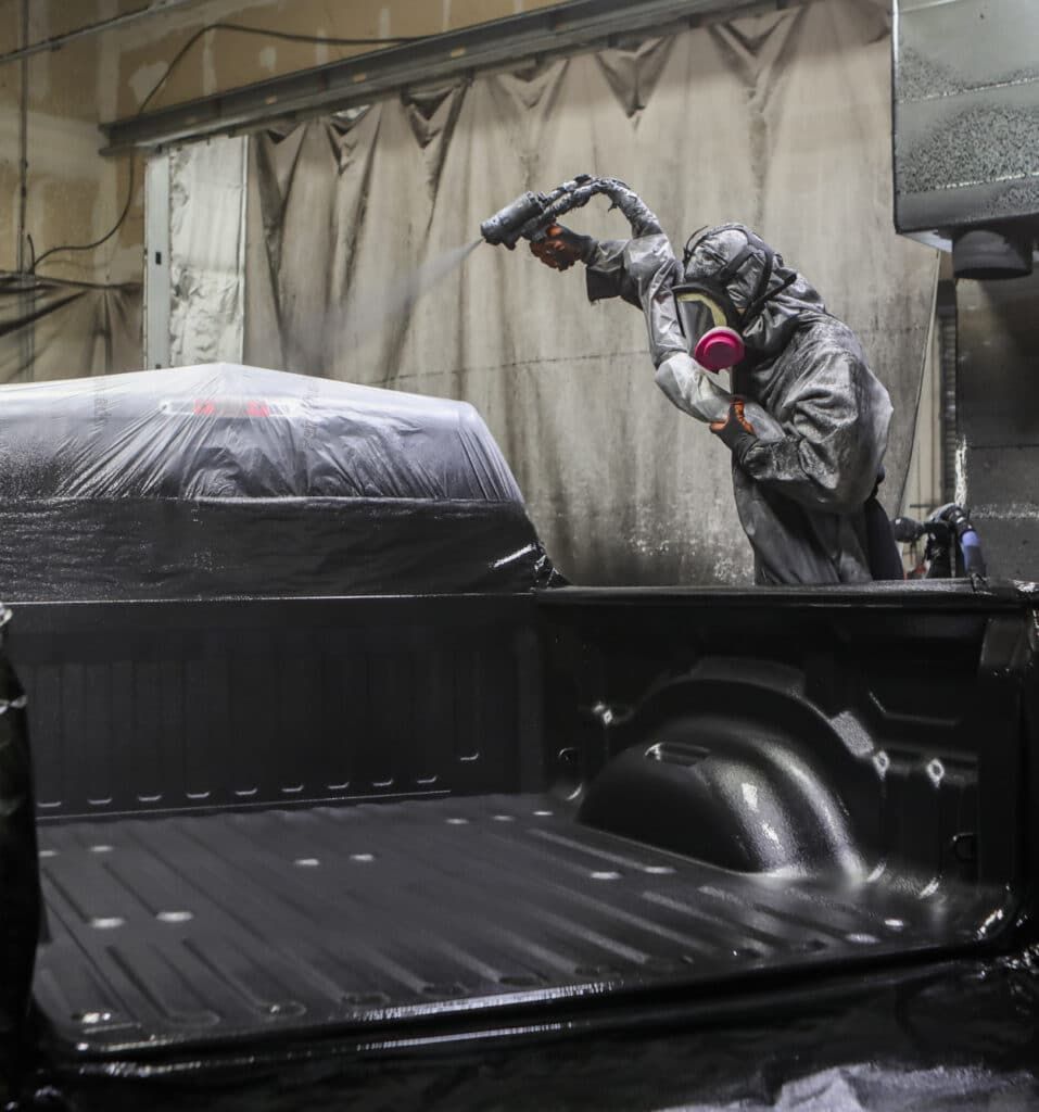 Person in protective suit spraying a truck bed with a dark coating in a workshop.