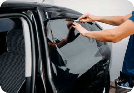 Person applying tint to a black car window, using a blade to cut the film.