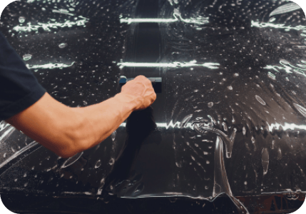Person applying window tint, using a tool to smooth the film over a dark surface covered in bubbles.