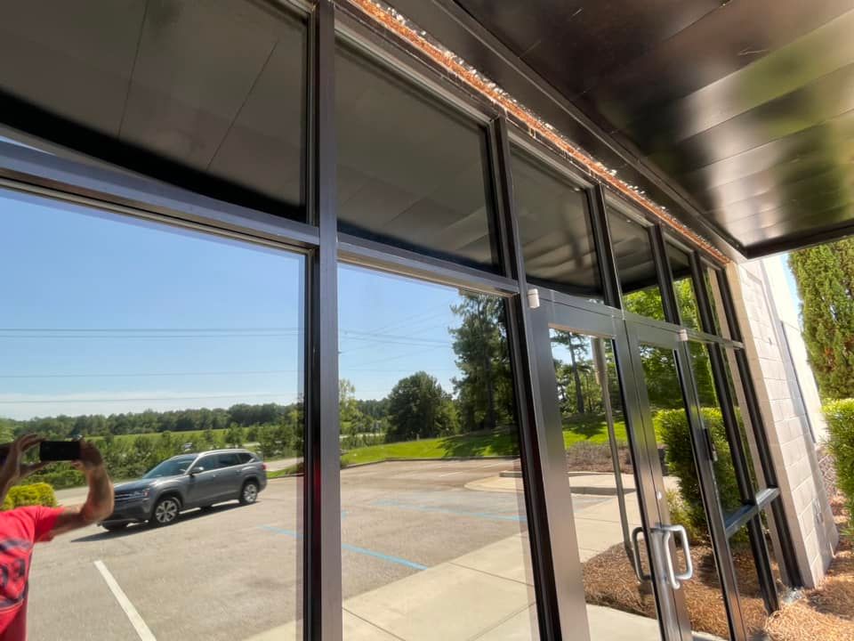 Exterior of a building with large reflective windows; a person takes a photo of a car and landscape reflected in the glass.
