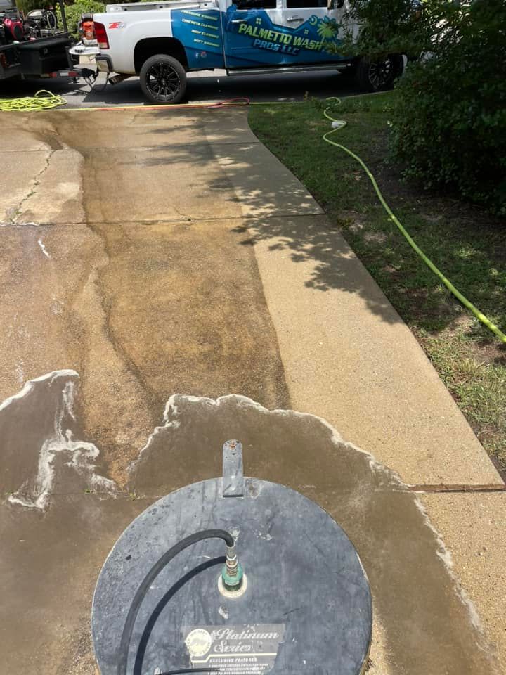 A power washer cleaning a concrete driveway, next to grass. A truck is parked nearby.