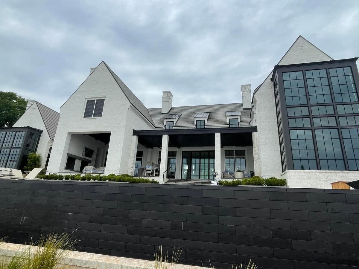 White brick house with black windows and retaining wall, cloudy sky.