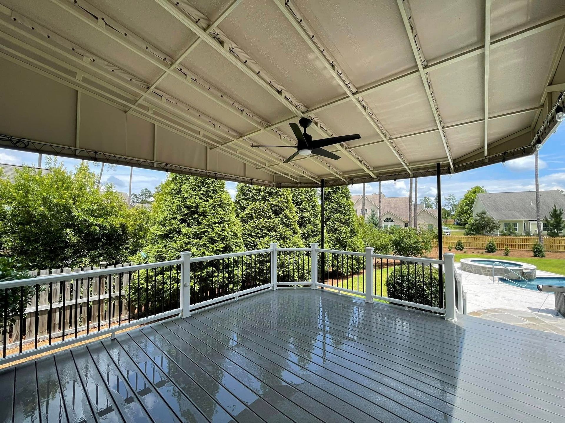 Covered deck with gray flooring, black railing, and a ceiling fan overlooking a backyard with trees and a pool.