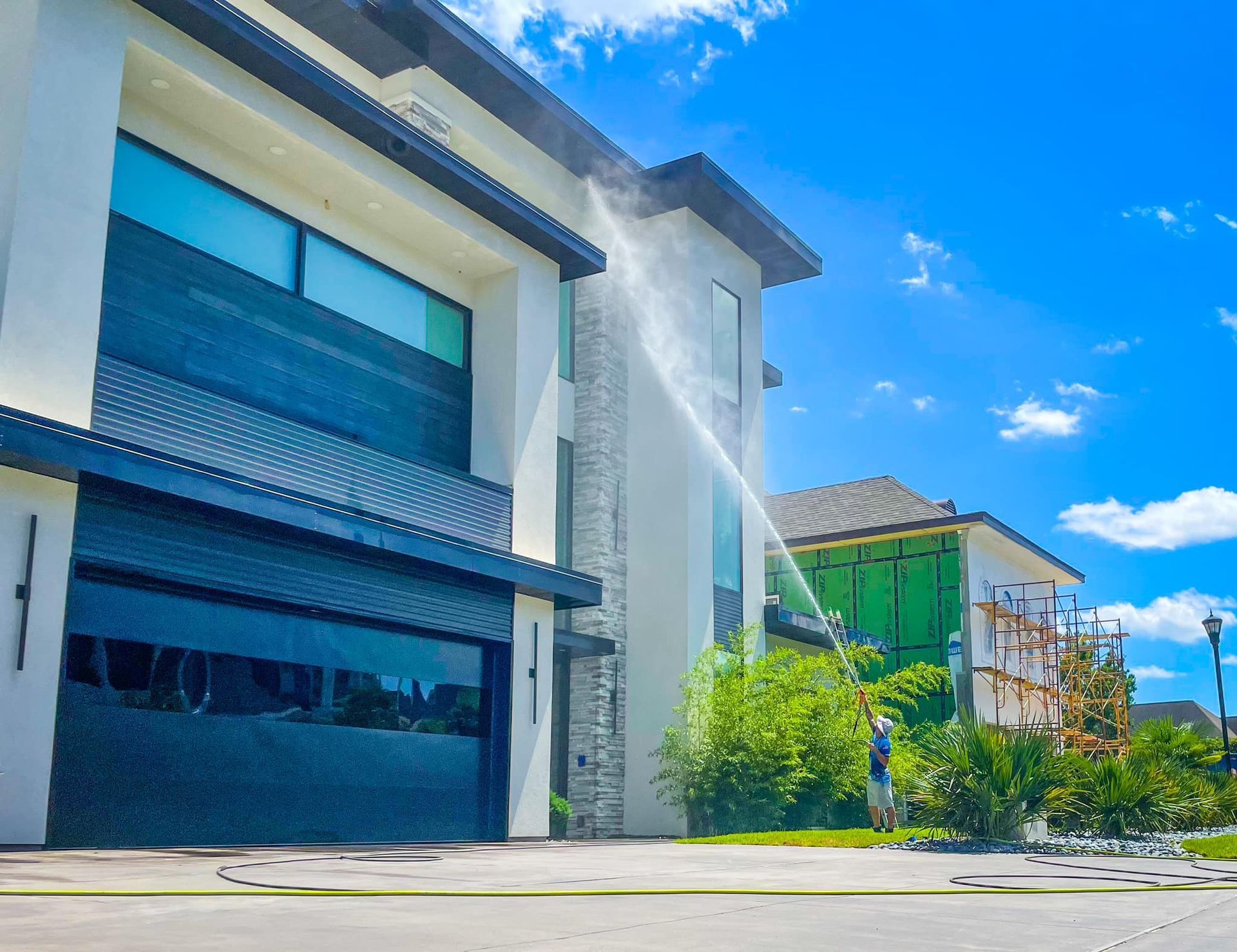 Man washing a modern two-story house with water. Blue sky. Another house in background.