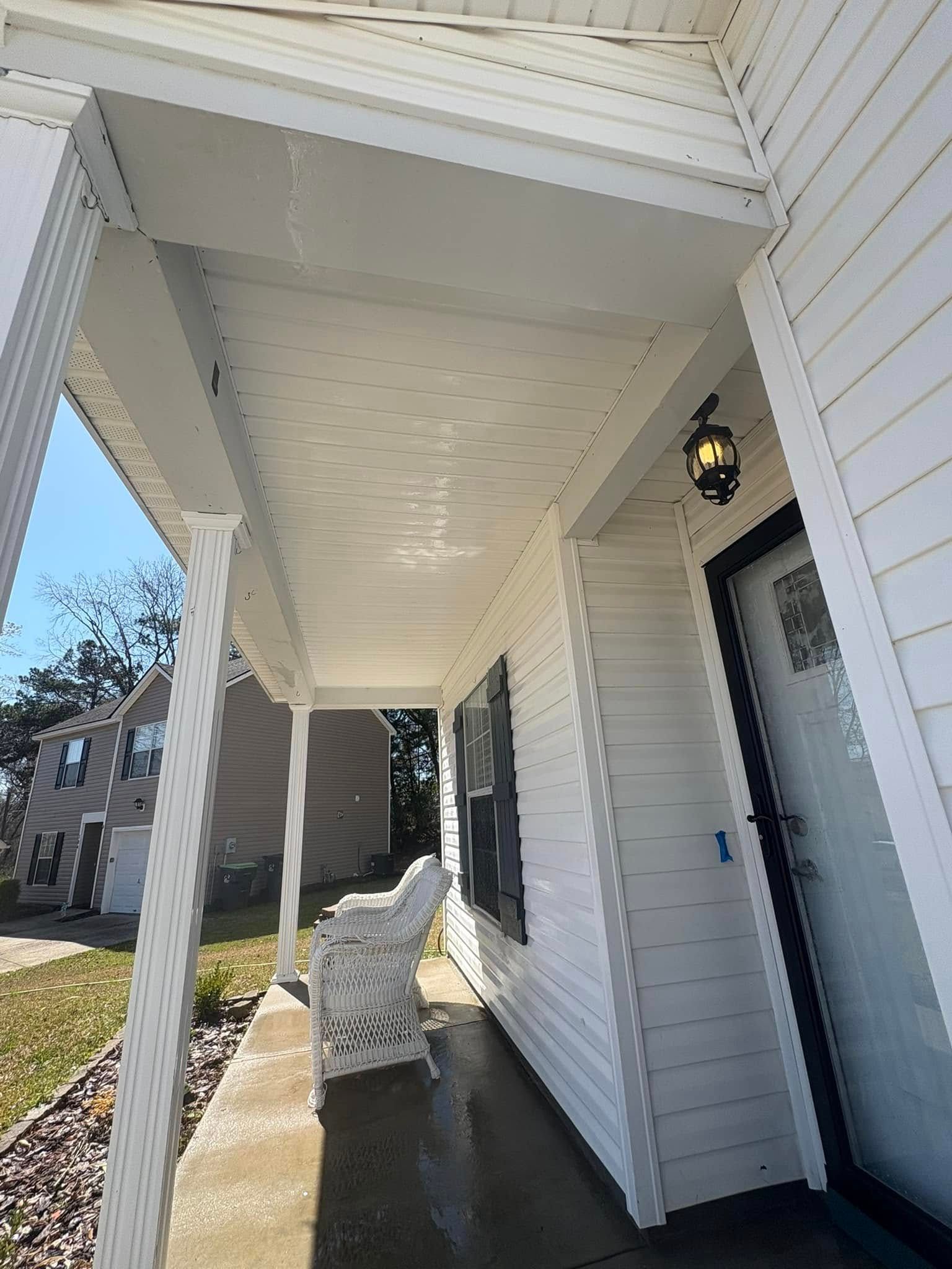 White front porch with a chair, next to a house with a black door and a lamp.