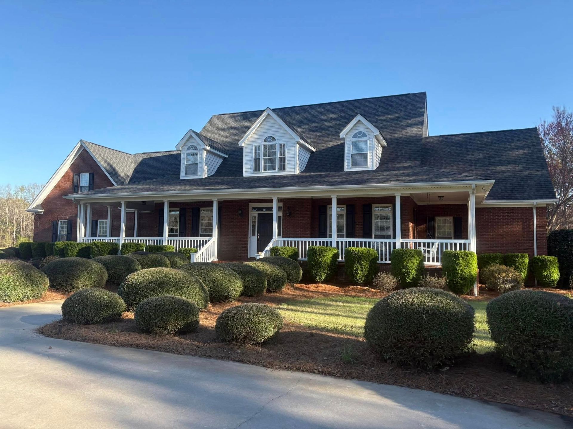 A two-story brown house with a wrap-around porch and manicured bushes under a blue sky.