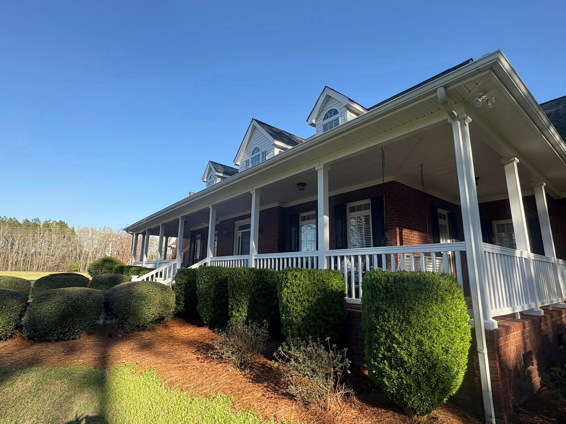 Brick house with white porch, trim, and shrubs under a blue sky.