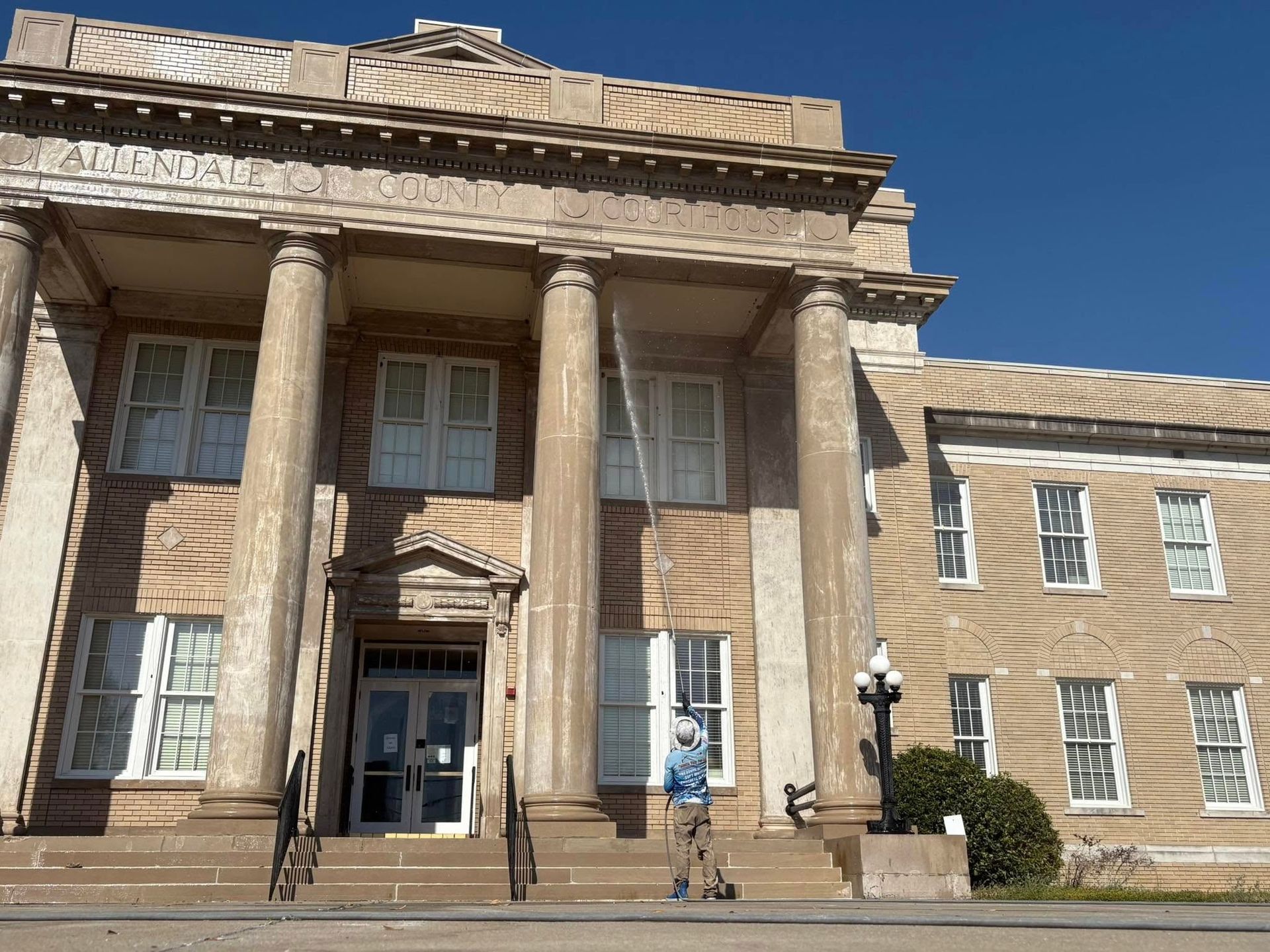 Person standing on steps of a classical-style building with columns and a sign. Bright blue sky.