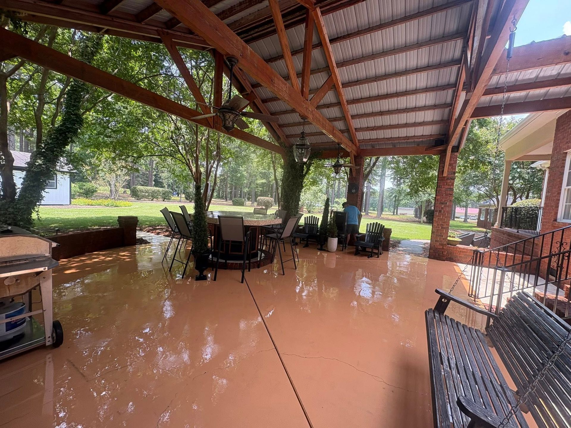 Covered patio with a brown floor, seating, and trees in the background.