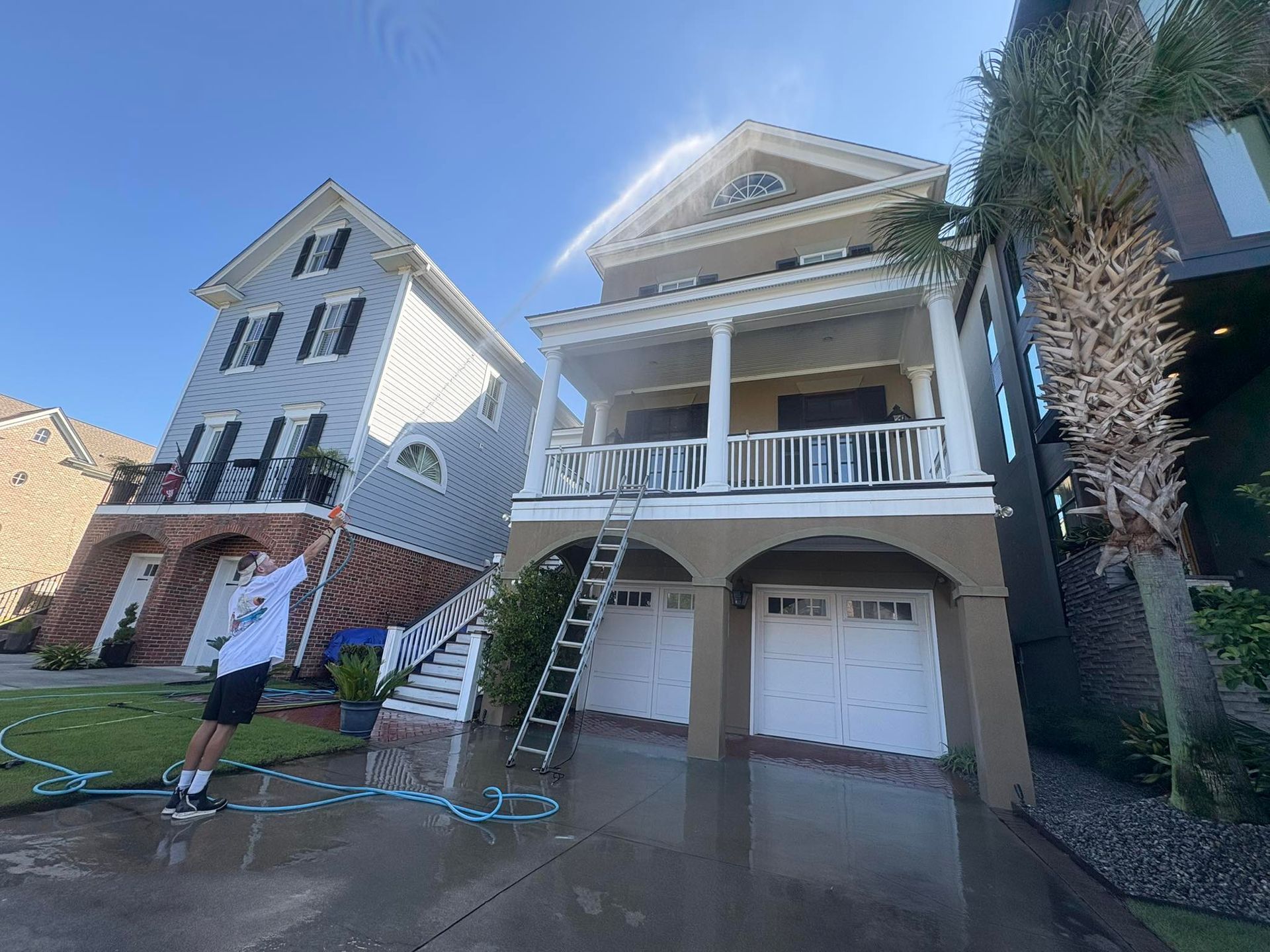 Person spraying water on two-story houses. One house is gray, the other beige, with a ladder leaning against it. Sunny day.