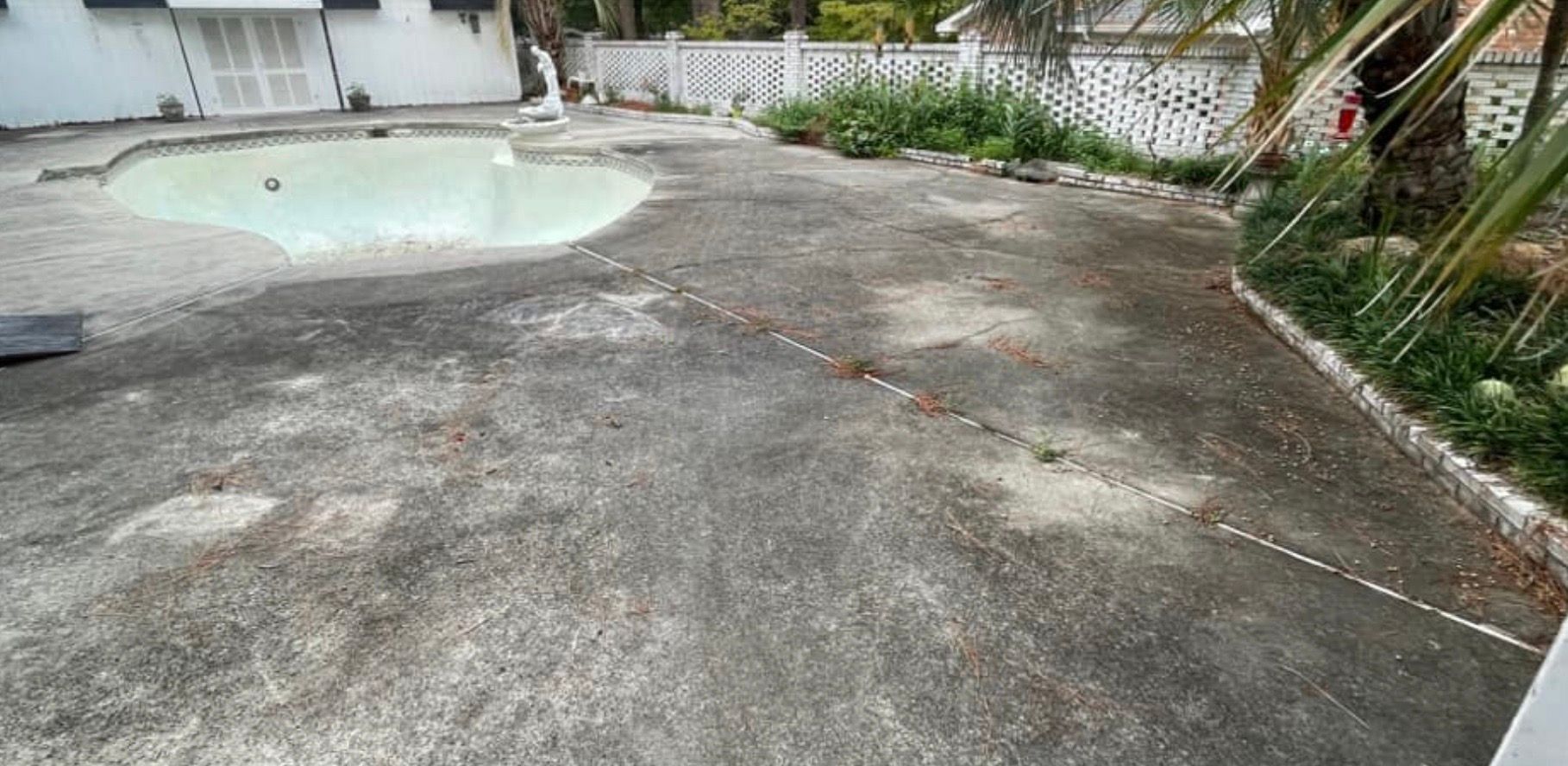 Exterior view of a weathered concrete area with a drained pool, surrounded by plants and a white fence.