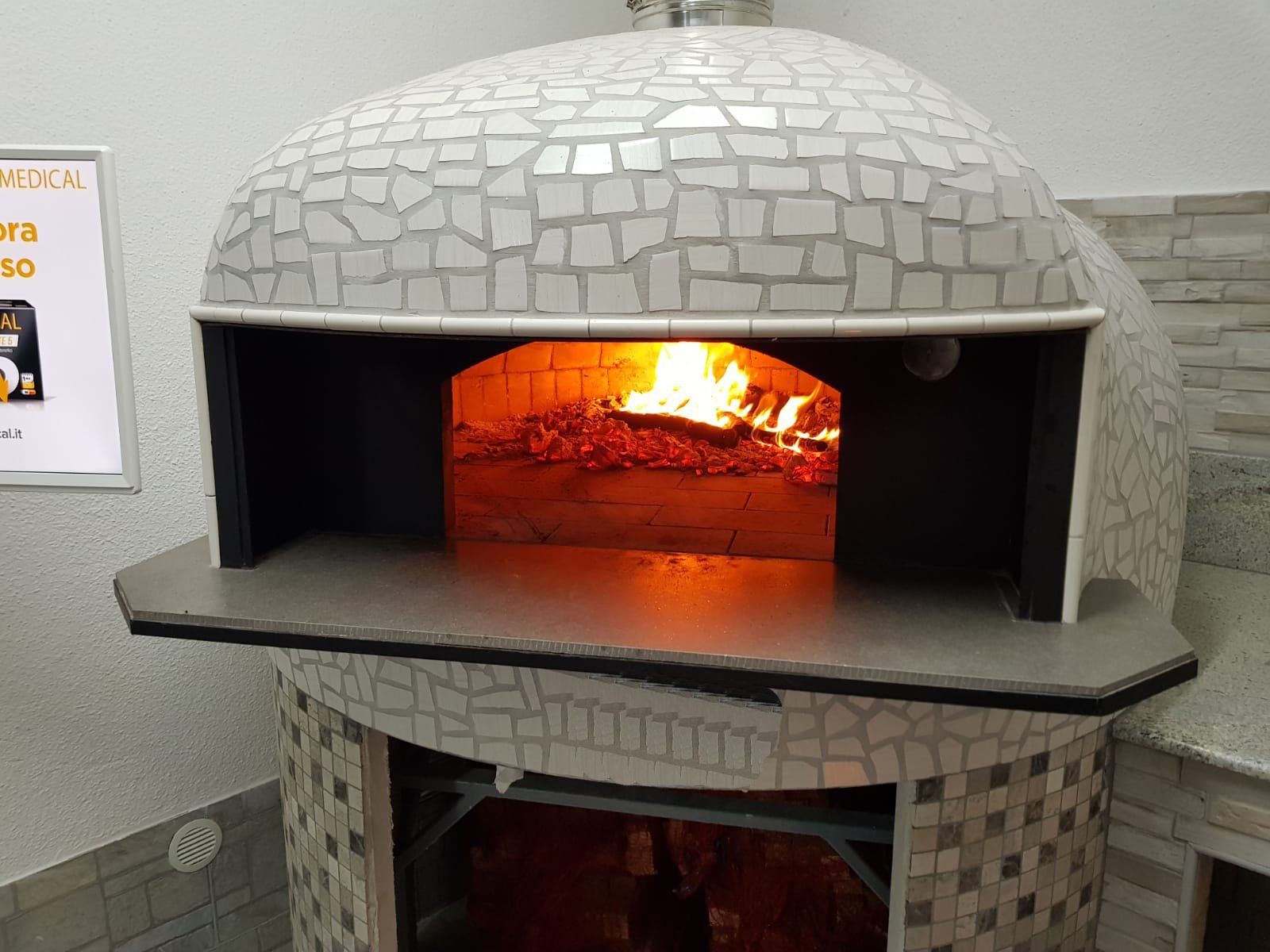 Forno a legna per pizza, vista interna con fiamme e braci, esterno in piastrelle a mosaico grigie e bianche.