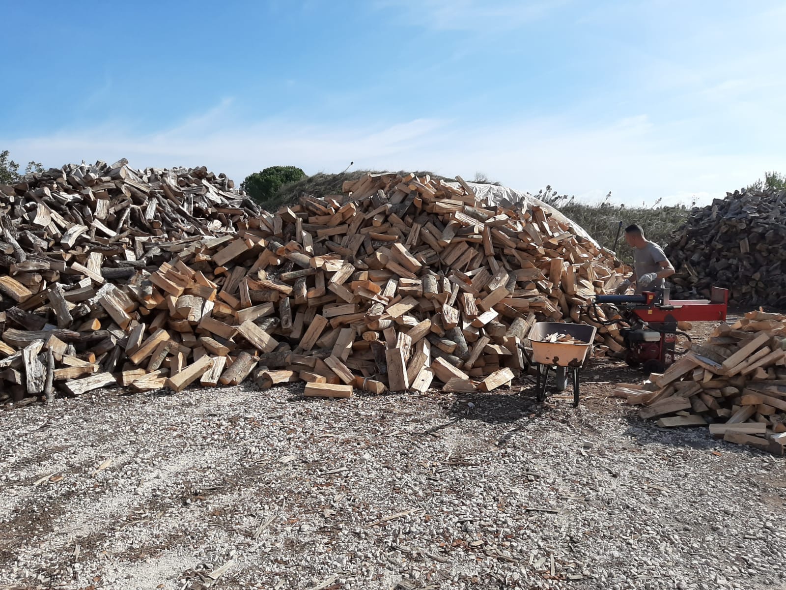 Un uomo accatasta legna da ardere spaccata all'aperto su un terreno ghiaioso sotto un cielo azzurro e terso.