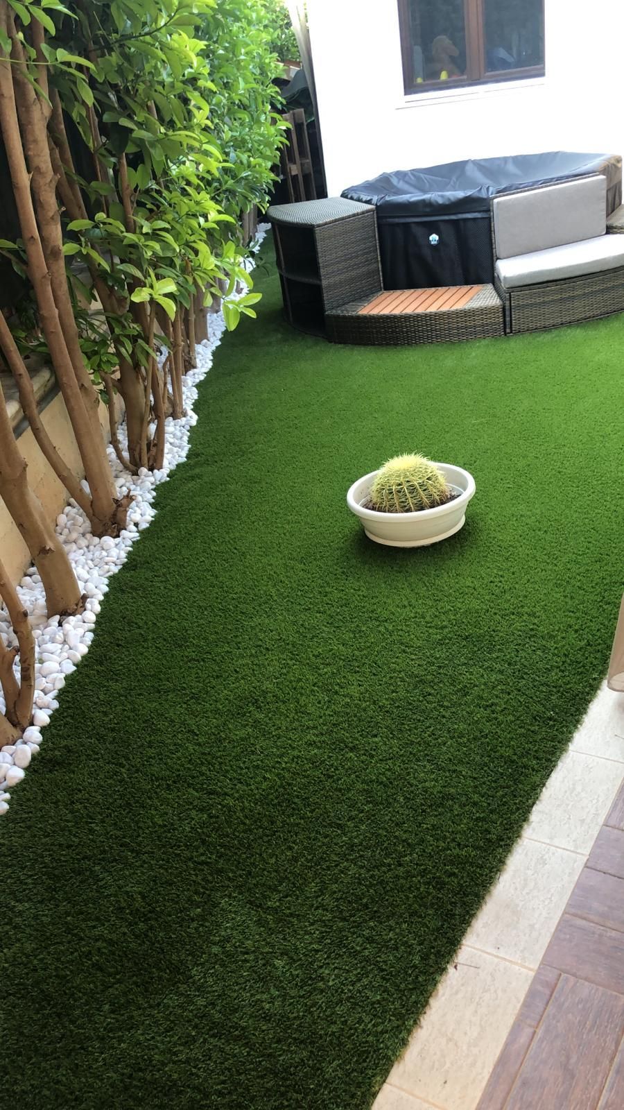 Un rigoglioso prato artificiale verde con pietre bianche, una pianta in vaso e una vasca idromassaggio in un ambiente esterno.