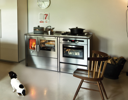 Una cucina con fornelli e forno in acciaio inox. Un cagnolino è seduto sul pavimento e una sedia è posizionata davanti ai fornelli.