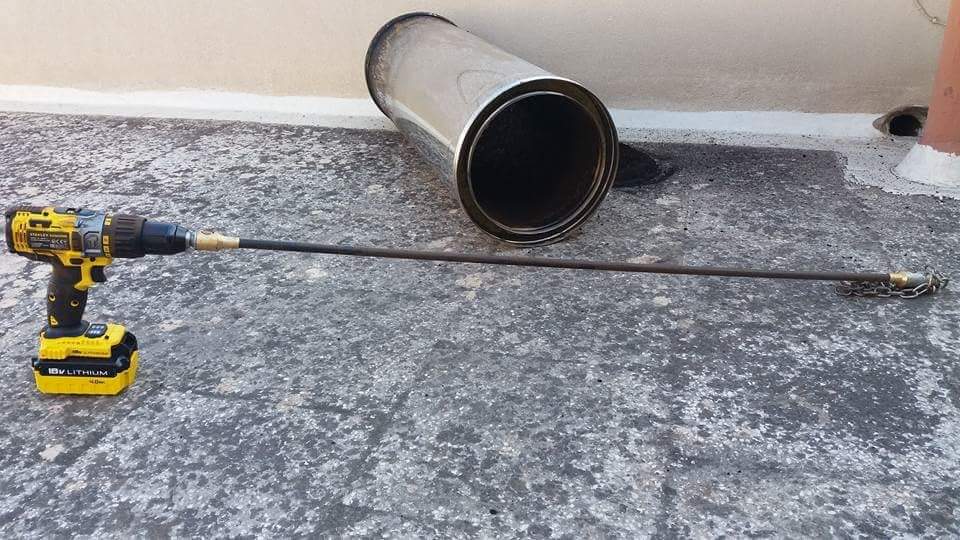 Un trapano giallo con una spazzola per la pulizia del camino e un tubo di metallo su una superficie di cemento.