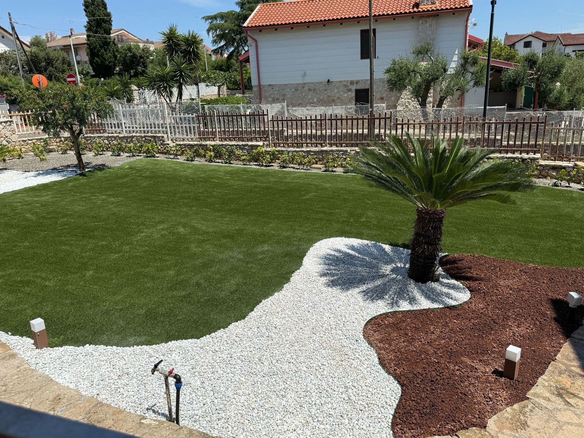 Un giardino con un sentiero in pietra, erba sintetica e una piccola palma. Sullo sfondo, pacciame marrone e una recinzione.