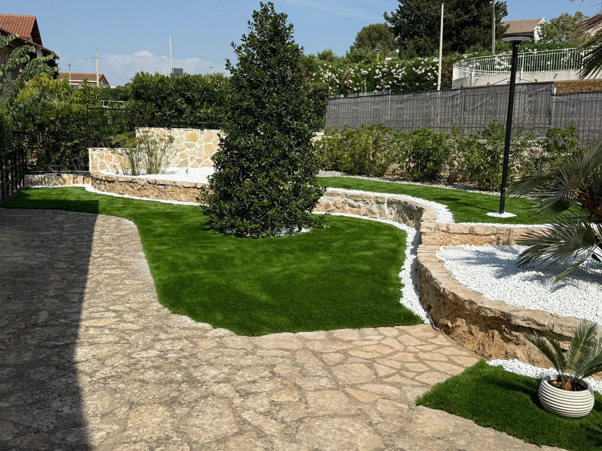 Un sentiero in pietra attraversa un giardino con erba verde, muri in pietra, pietre bianche e un piccolo albero.