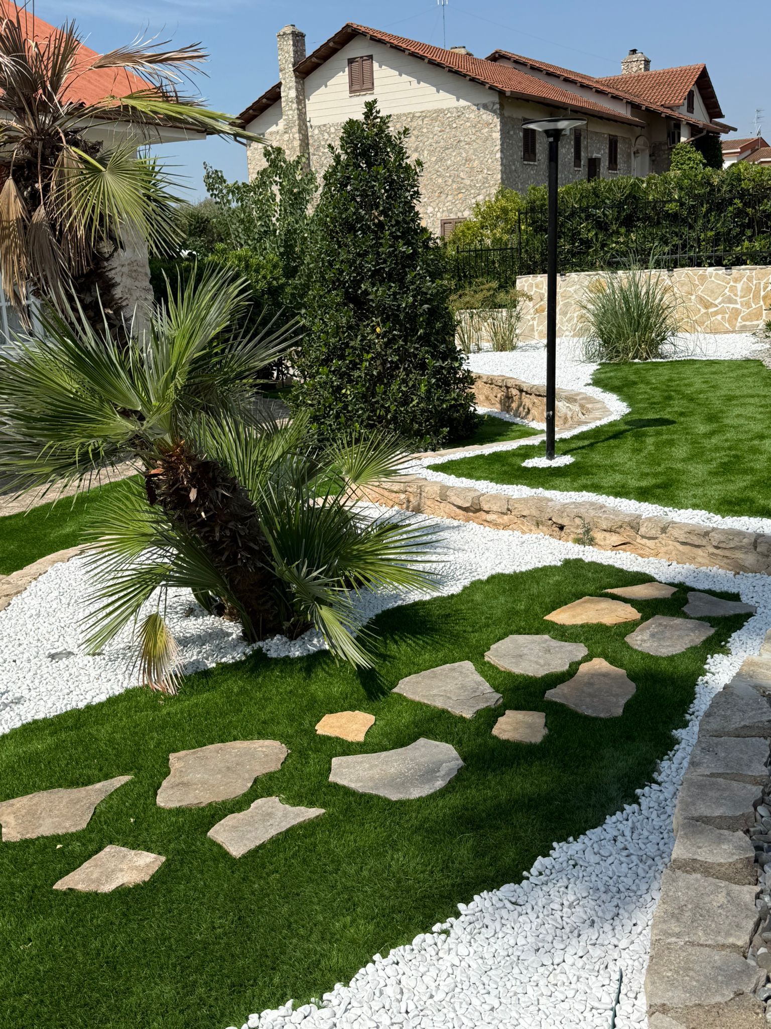 Un giardino paesaggistico con sentiero in pietra, erba verde, ciottoli bianchi e una palma di fronte a una casa in mattoni.