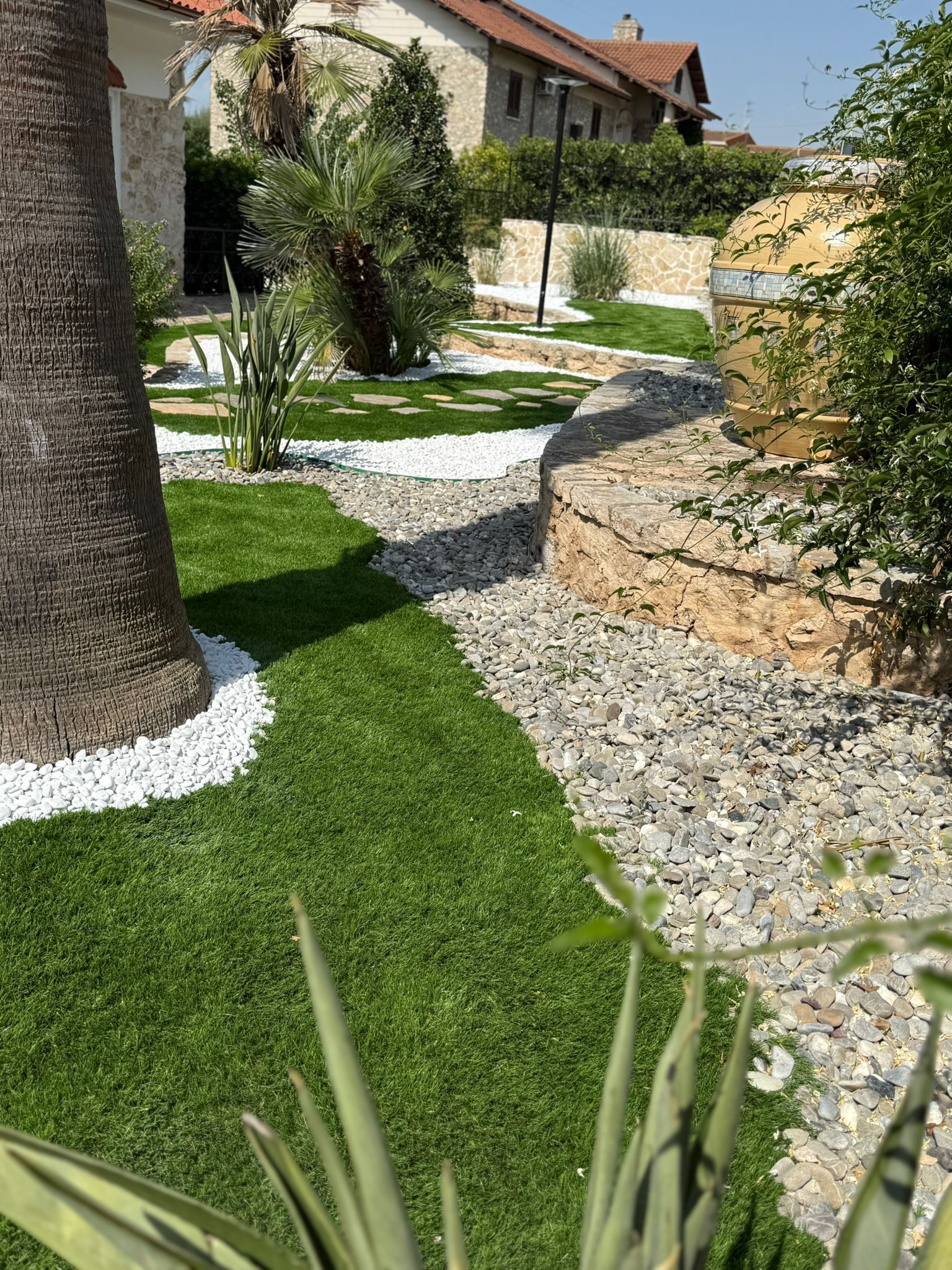 Un rigoglioso prato verde, rocce bianche e un sentiero di ghiaia che si snoda attraverso un giardino paesaggistico con un muro di pietra beige.