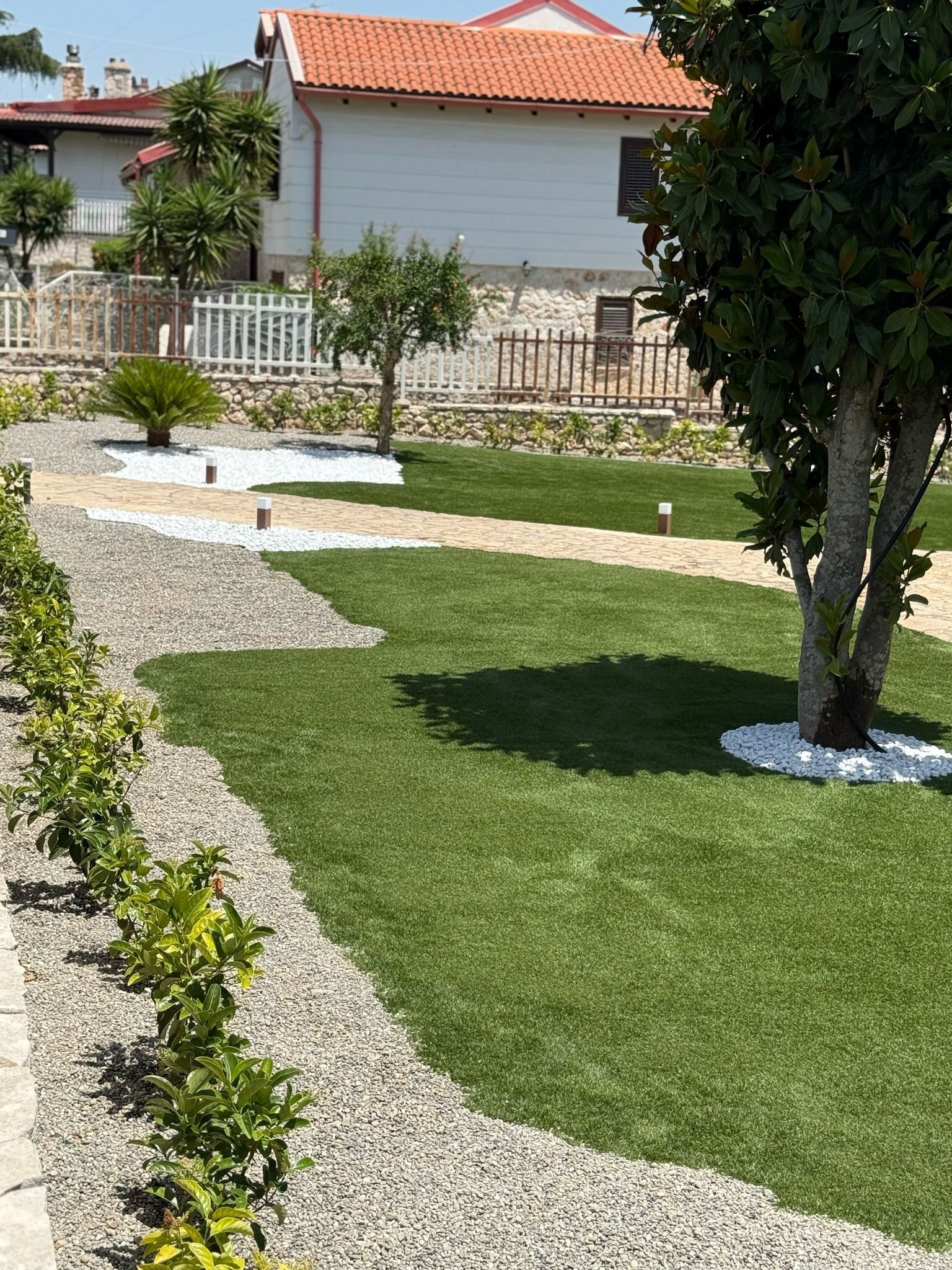Prato verde con alberi e bordi di ghiaia di fronte a un edificio bianco con il tetto rosso.