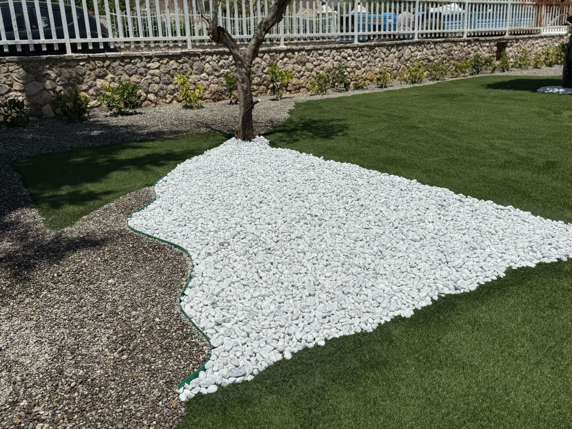 Un cortile con ghiaia bianca attorno a un albero, accanto a erba artificiale verde e ghiaia marrone.