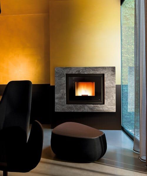 Camino con cornice in pietra su parete gialla, sedia nera e pouf vicino alla grande finestra.