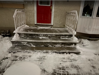 Snow-covered front steps leading to a red door, with icy railings and surrounding snow.