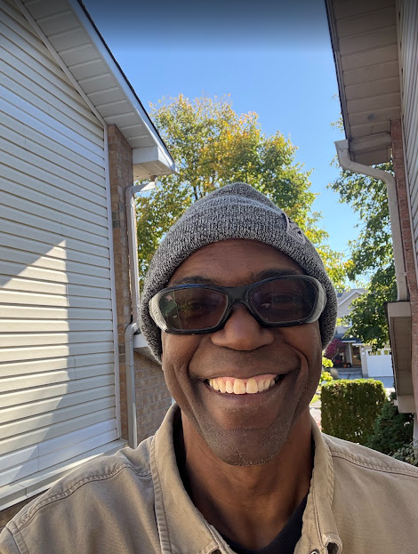 Man smiling, wearing a gray beanie and sunglasses, standing between two buildings on a sunny day.