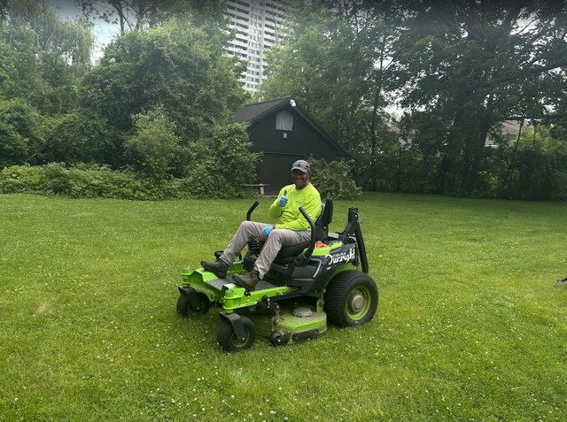 Man on green riding lawnmower on a grassy lawn with a small building in the background.