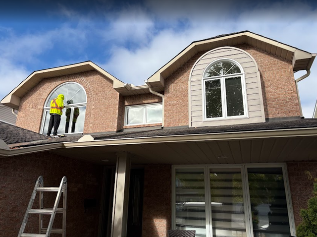 Person in yellow jacket near window on house, ladder in view, blue sky background.