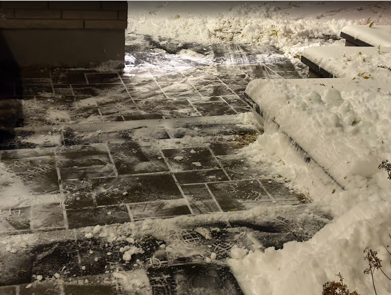 Snow-covered brick walkway and steps at night. Partially cleared path and piled snow on sides.