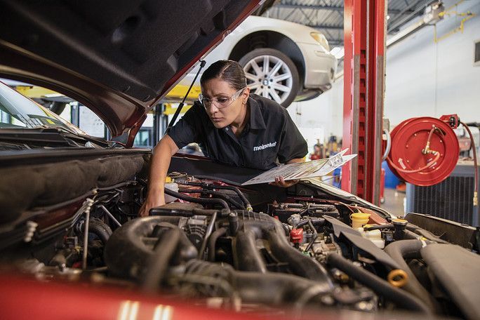 A woman is looking under the hood of a car in a garage. | Meineke Car Care Center