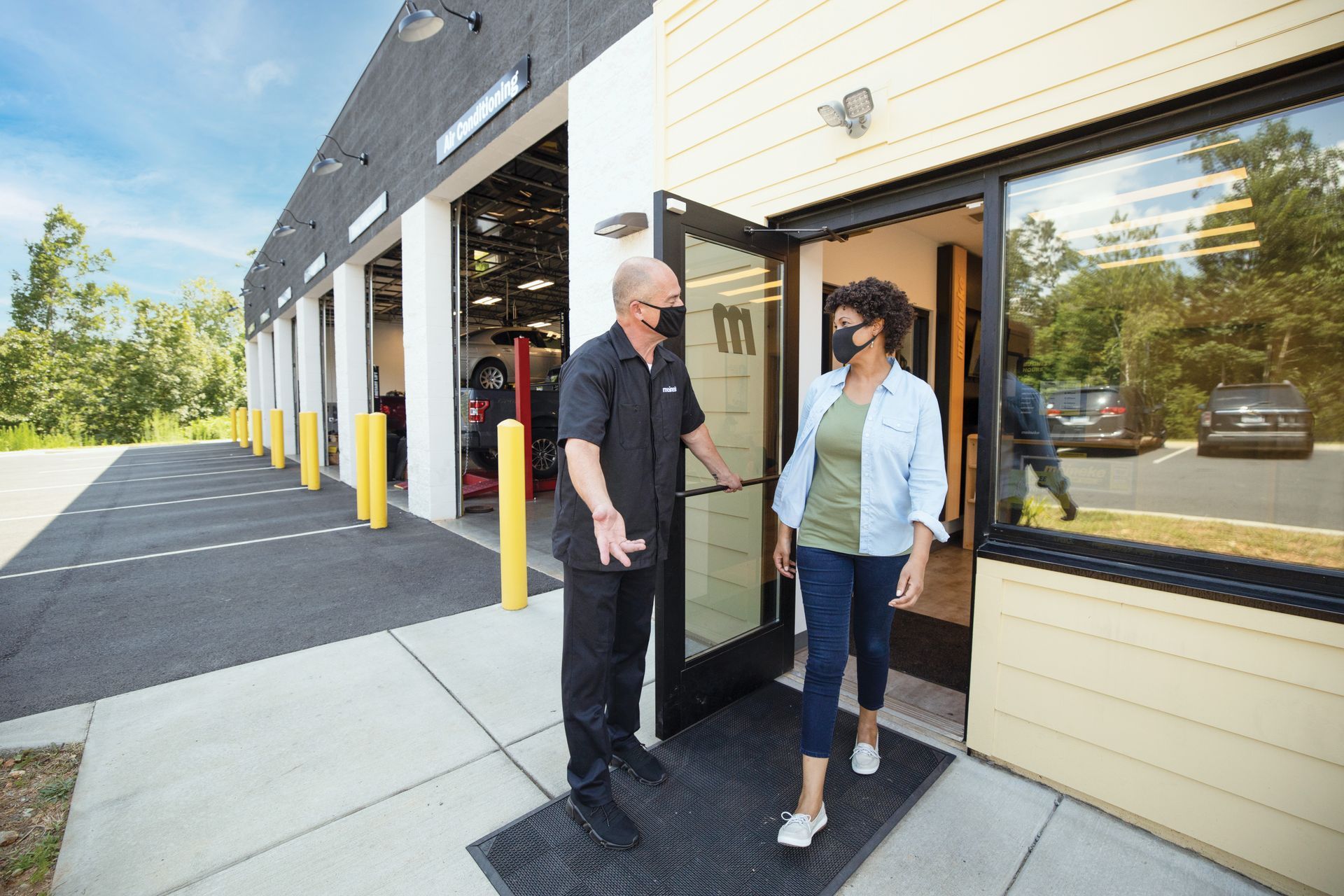 A man and a woman wearing face masks are standing outside of a garage. | Meineke Car Care Center