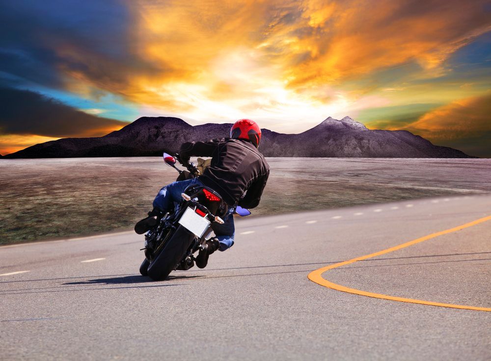 Motociclista inclinato in curva su una strada con sfondo montano, sotto un tramonto arancione.